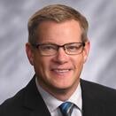 Paul smiles against a gray background, wearing a black suit jacket with a blue tie. Paul has blonde hair and wears glasses.