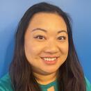 Lillian smiles wearing a green shirt in front of a blue wall. She has long dark brown hair and dark brown eyes. 