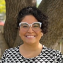 Trin wears a geometric blouse and stands in front of a tree. She has a short black bob and wears white framed glasses.