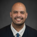 Dr. Sadak's face fills the frame, he is bald with slight facial hair and is smiling. He wears a black sweater over a white button up and tie.