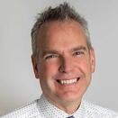 Dr. Shreve smiles at the camera. He has spiky grey hair and wears a white button up with a dark tie.