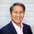 A man smiling. He is wearing a suit with a light pink button down.