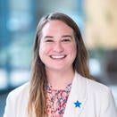 A woman smiling, wearing a white jacket with long straight brown hair.