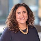 A woman with curly dark brown hair, wearing a dark sweater. 