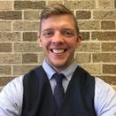 Timothy in a black vest and tie smiling in front of a brick wall