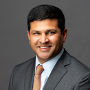 Vik Bhatia smiling and wearing a blue s hirt, orange and blue tie, and grey suit jacket