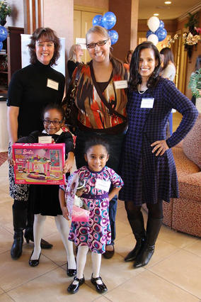 Three adults in elegant clothing stand with arms around each other. In front of them, two children wearing party dresses, white tights, and black patent shoes happily show off boxes containing new toys.