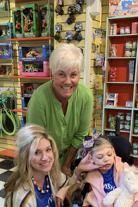 A woman with short white hair wearing a lime green shirt leans down next to a child in a wheelchair. The child is wearing a pink polka-dot jacket with a matching hairbow. Kneeling down beside the child is a woman with long blond hair. The background is a toy shop. 