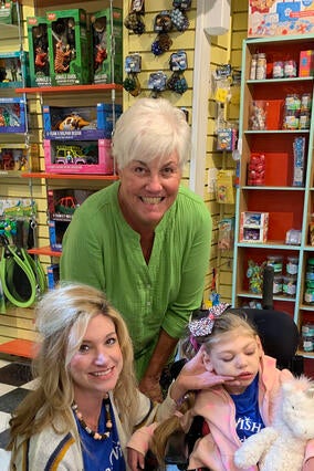 A woman with short white hair wearing a lime green shirt leans down next to a child in a wheelchair. The child is wearing a pink polka-dot jacket with a matching hairbow. Kneeling down beside the child is a woman with long blond hair. The background is a toy shop. 