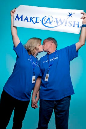 Charlie & Lori holding a Make-A-Wish banner and wearing matching Make-A-Wish t-shirts kissing each other