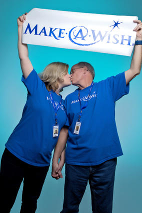 Charlie & Lori holding a Make-A-Wish banner and wearing matching Make-A-Wish t-shirts kissing each other 