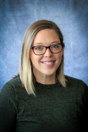 Kristen smiles in front of blue background. Wearing black sweater and glasses, mid length blonde hair.