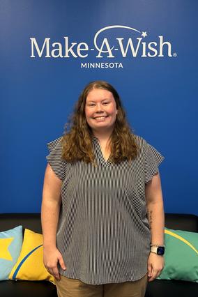 Maddi stands smiling in a stripped black and white shirt with red curly hair in the Make-A-Wish Minnesota office in front of a blue wall with the Make-A-Wish decal.