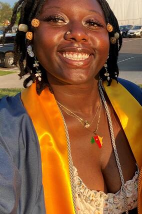 Nkobwa smiles outside wearing braids with beads and a white and orange top