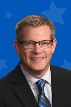 Paul wears a black suit coat, stripped blue and gray tie, white shirt and glasses. He smiles at the camera against a blue background with stars.
