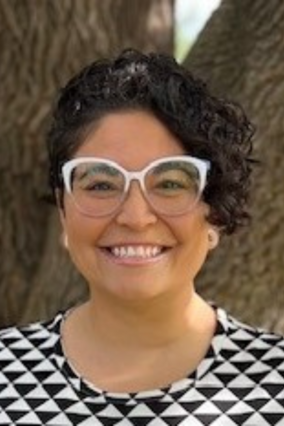Trin stands in front of a tree wearing a black and white geometric shirt. She has a short black bob and wears white framed glasses.