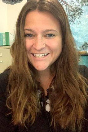 Megan smiles at camera, she has long brown hair and wears a black shirt with a large necklace.