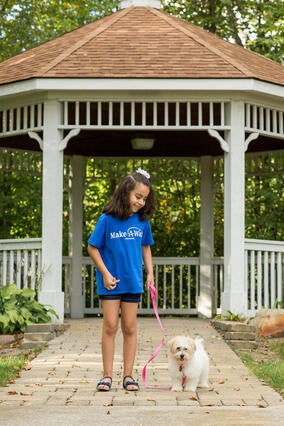 A 7-year-old child with shoulder-length curly dark hair looks down with joy at a fluffy white puppy, whose hot pink leash the child is holding. The child is wearing a royal blue Make-A-Wish t-shirt, jean shorts, blue sandals, and a white hair bow.The two are walking along a paved path that leads away from a white gazebo.