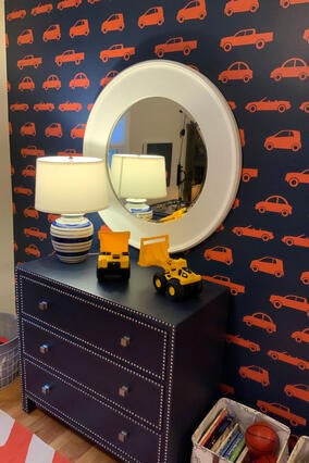 A wall in a bedroom painted navy blue and patterned with small orange cars and trucks. In front of the wall is a navy blue dresser accented with silver metal circles. On the top of the dresser is a blue and white lamp, a white mirror, and two plastic toy trucks. Other bins of toys and books can be seen on the floor nearby.