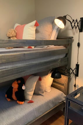 A set of gray bunk beds made up with knitted gray blankets and pillows. There are also several white and orange accent pillows, a stuffed gorilla, and a stuffed bear. Next to the beds sits a navy blue end table accented with round silver studs. 