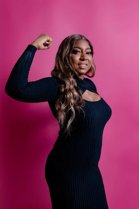 Jakeina today wearing a black dress and holding her right arm up to show a muscle