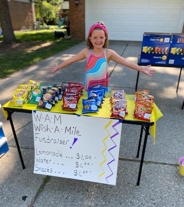 A child in a hot pink headband and brightly colored dress stands with arms thrown out wide behind a snack stand fundraiser. In front of the child, a table covered with a bright yellow plastic tablecloth is filled with individual variety bags of potato chips. A sign taped to the front of the table reads "WAM (Wish-A-Mile) Fundraiser" and advertises lemonade, water, and snacks for sale.