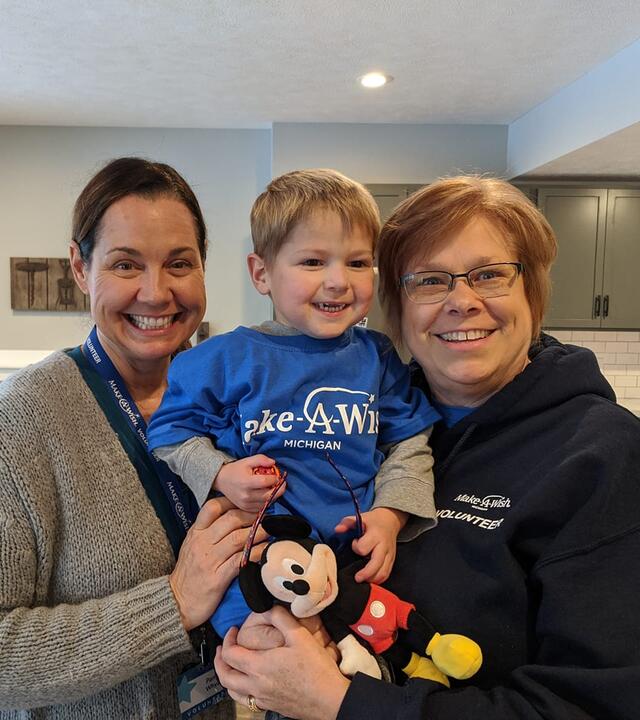 A young child with short blond hair is wearing a royal blue Make-A-wish t-shirt over a long-sleeved gray shirt and clutching a stuffed Mickey Mouse toy as two wish-granting volunteers hug him during a wish discovery meeting.