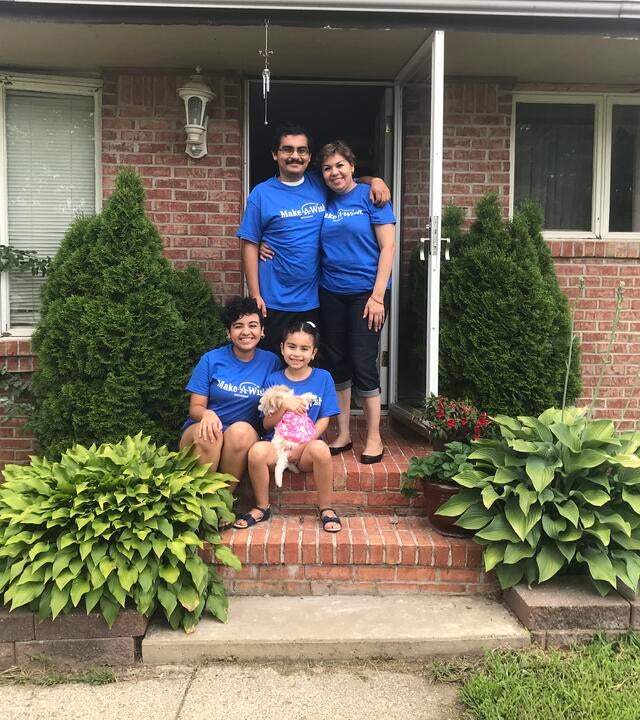 A family of two adults and two children, all wearing royal blue Make-A-Wish Michigan t-shirts, poses for a photo in front of a red brick home surrounded by greenery. The youngest child holds their wish, a fluffy white puppy.