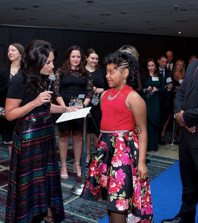 A wish child in a red and black floral patterned dress speaks with a news anchor during a formal gala.