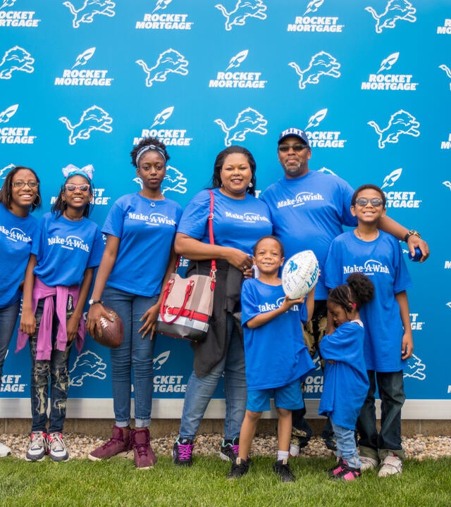 A family of two adults and 6 children, all wearing royal blue Make-A-Wish Michigan t-shirts, pose for a picture in front of a blue background with the Detroit Lions and Rocket Mortgage logos. The wish kid in the front of the group is holding up a signed football.