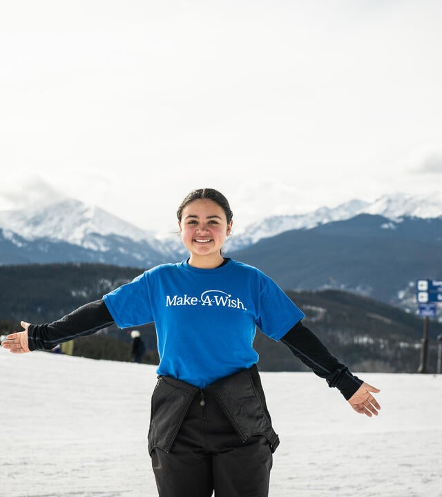 Wish kid Antonia standing with open arms on ski mountain wearing Make-A-Wish shirt