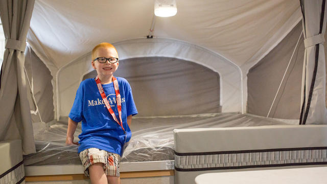 A child with short red hair and black glasses sits on the bed inside a camper. The child is wearing a royal blue Make-A-Wish t-shirt, tan plaid shorts, and a red lanyard with a large tag that says VIP.