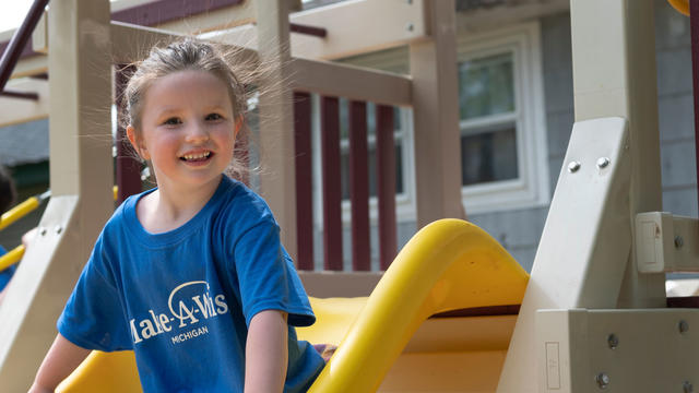 A young child in a royal blue Make-A-Wish t-shirt and multicolored plaid skirt sits at the top of a yellow playground slide with a wide smile. 