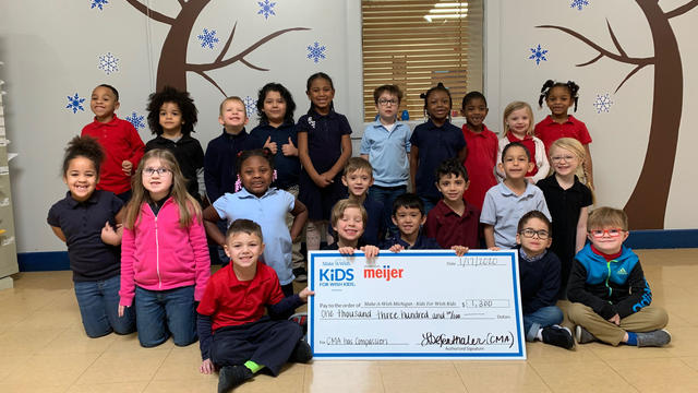 A group of 22 elementary school students gather for a group photo. The children in the front hold an oversized check made out for $1,300 with the Kids For Wish Kids and Meijer logos. Behind them is a white wall decorated with images of leafless trees and snowflakes.