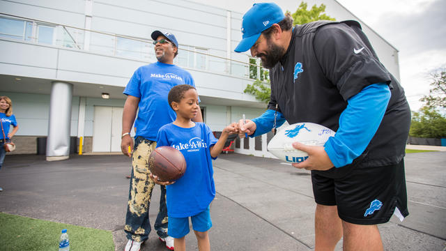 A child in a royal blue Make-A-Wish t-shirt and jean shorts holds a football in one hand and uses the other hand to fistbump an adult in Detroit Lions clothing who is signing a second football. 