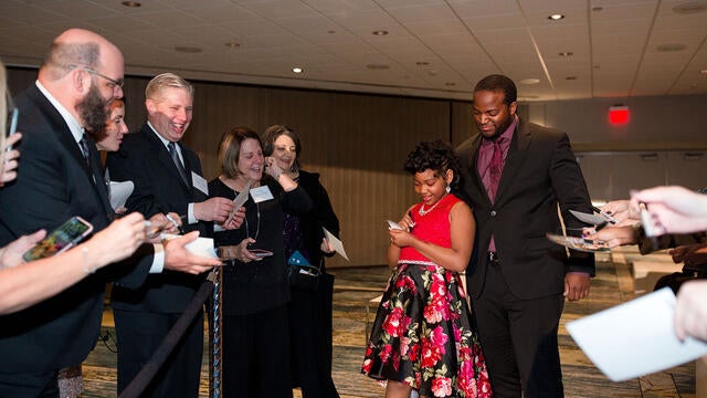 Wearing a red and black dress patterned in bright florals, a child with dark curly hair worn in a fancy updo signs an autograph on a red carpet leading into a ballroom. A crowd of admirers stands on either side, taking photos and requesting autographs, while a tall adult in a black suit stands behind the child, playing the part of bodyguard.