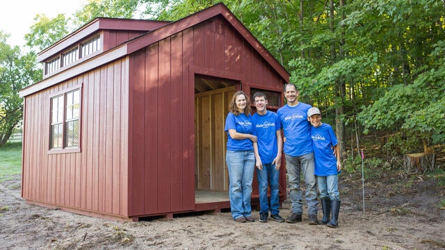 A family of four wearing royal blue Make-A-Wish t-shirts stands in front of the open door to a red outdoor shed.