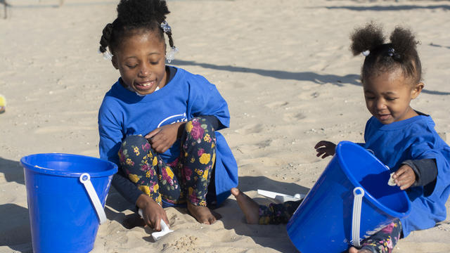 Two children in royal blue Make-A-Wish t-shirts play happily on a beach of white sand. Each child has a royal blue bucket and shovel.