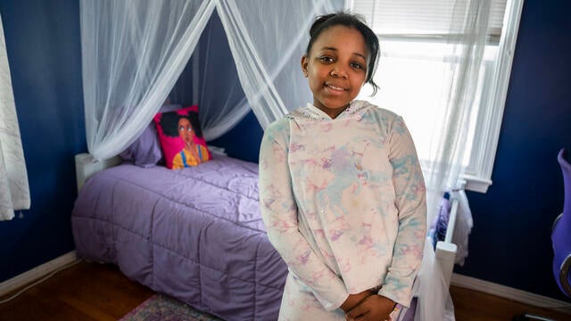 A child wearing a pastel tie-dyed hoodie with a unicorn design and matching pants stands with hands clasped in front of a bed decorated with sheet white curtains and a purple bedspread.
