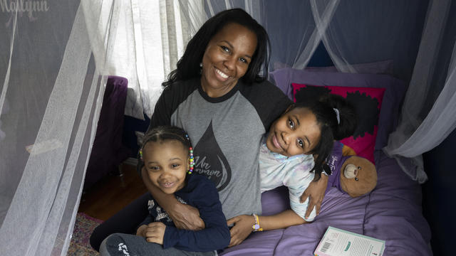 An adult and two children cuddle on a bed decorated with sheer white curtains, a purple bedspread, and pink and purple accent pillows.