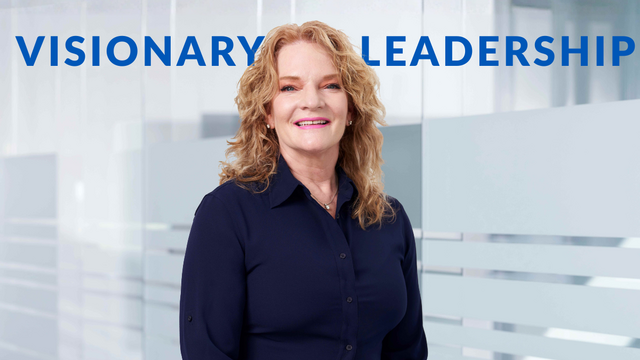Smiling woman with wavy blonde hair in a blue collared shirt, against a blurred glass-walled background with "VISIONARY LEADERSHIP" in blue text above.