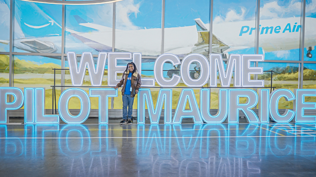A Black child with locks stands smiling in front of large, glowing neon letters that spell "WELCOME PILOT MAURICE" with an airplane with "Prime Air" on its side visible outside, the floor reflecting the sign's neon glow. He wears a brown leather jacket, a blue t-shirt with a logo and jeans.