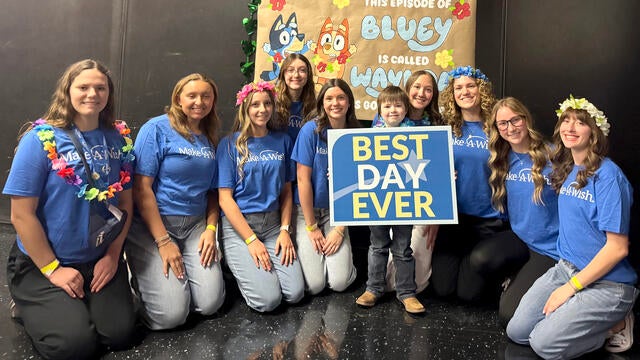 Group of Make-A-Wish volunteers in blue shirts pose with a child holding a “Best Day Ever” sign in front of a handmade Bluey-themed backdrop for a wish event.